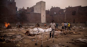 Fotografia de campo indiano onde estão sendo realizadas piras funerárias para mortos da pandemia, com homem trazendo novo cadáver - Getty Images