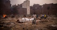Fotografia de campo indiano onde estão sendo realizadas piras funerárias para mortos da pandemia, com homem trazendo novo cadáver - Getty Images