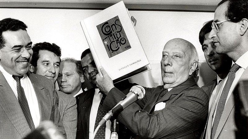 Ulysses Guimarães segurando uma cópia da Constituição de 1988 - Agência Brasil via Wikimedia Commons